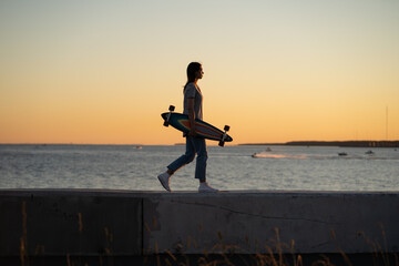 Lifestyle and freedom concept: young girl with longboard walking over sunset view. Silhouette of trendy teen or student female skater with skateboard at riverside pavement. Woman walk along river © DimaBerlin