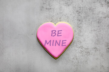 A heart shaped sugar cookie with pink royal icing and Be Mine text on it.