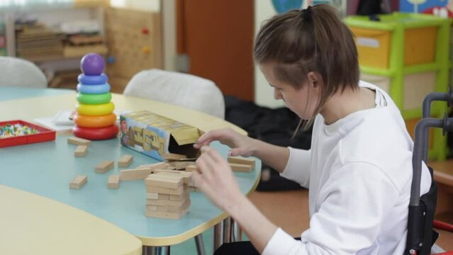 a girl with disabilities builds a tower of wooden cubes, sitting at a table in a special chair