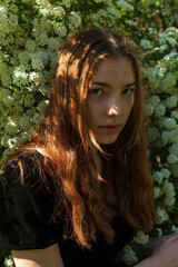 vertical photo of a beautiful red-haired woman sitting near a bush of white flowers looking pleasantly to the side, illuminated by the rays of the setting sun