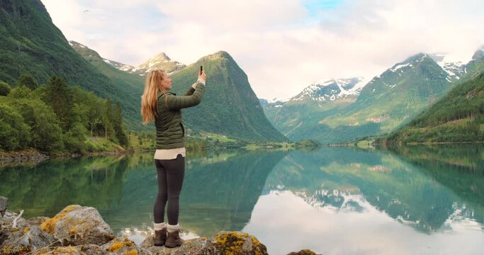 4k video footage of a young woman standing and using her cellphone to photograph the lake in Stryn