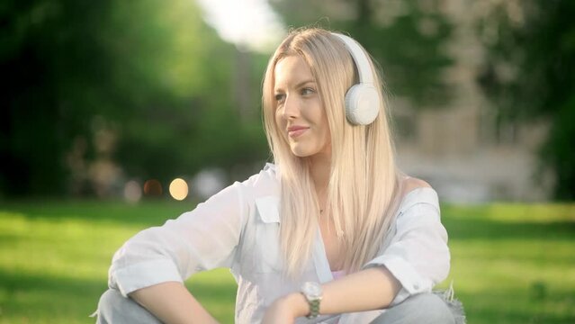 Young incredibly beautiful blonde woman in jeans and a shirt smiling while sitting on grass in a park listening to music on earphones