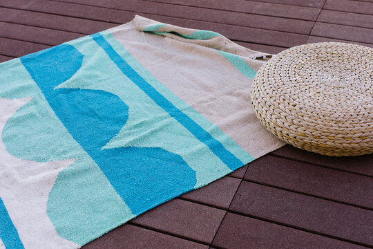 Blue and teal blanket sits on deck outside, next to woven poof
