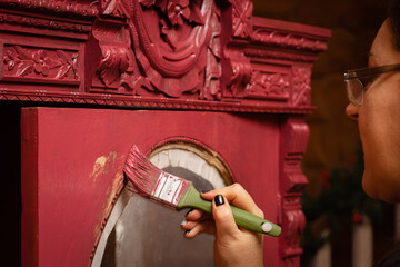 Woman carefully aging carved ornaments of antique wooden cupboard in pink color with paint brush....