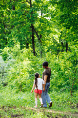 Back view of family walking in park forest around green trees, talking, having fun. Little cheerful daughter holding hand of middle-aged bearded man father. Summer activities, travelling. Vertical.