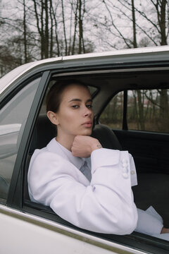 girl in a white suit with collected hair sits in a car and looks through a lowered window