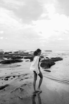 black and white photo of a girl in a black bikini and white shirt walking along the beach with large stones