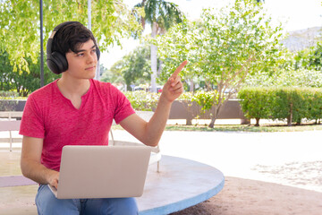 young man with laptop and wireless headphones in a park, looking at the camera and pointing a finger to the side. 