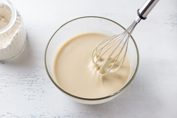 A glass bowl of dough for Finnish pancakes with a whisk and a jar of flour on a light gray background. Cooking delicious homemade food