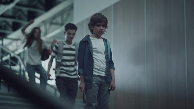 Sad little guy standing alone on school stairs. Pupils running downstairs.