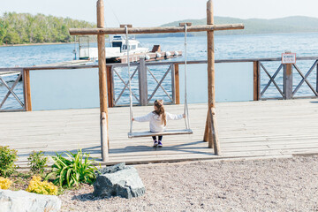A little girl swings on a wooden swing by the sea. Back view. Outdoor activities