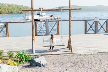 A little girl swings on a wooden swing by the sea. Back view. Outdoor activities