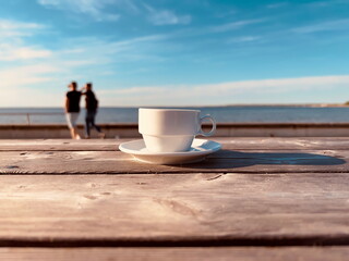 cup of coffee on wooden table in beach restaurant on horizon people walk and sea with blue sky