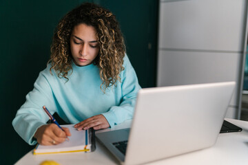 Focused woman writing in notepad sitting at laptop