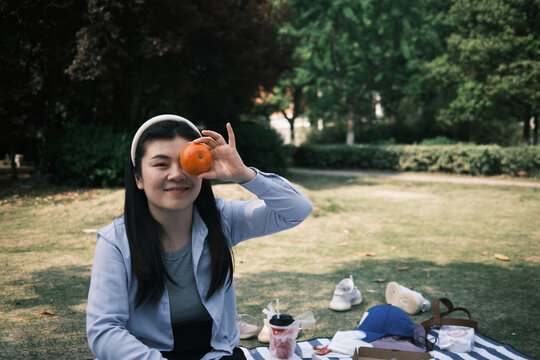 Cute Asian Woman Holding An Orange In Front Of Her Eye At A Picnic.