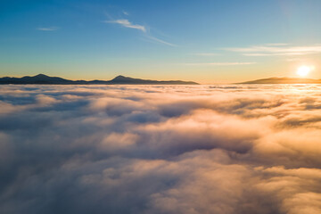 Aerial view of vibrant sunset over white dense clouds with distant dark mountains on horizon
