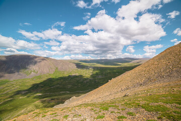 Scenic mountain landscape with green valley with river and mountain range with sunlight and shadows from clouds at changeable weather. Colorful alpine scenery with sunlit mountains and cloudy sky.