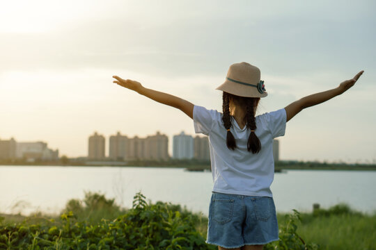 Asian kid girl wearing hat raising arms relaxing feeling happy looking at sunset on sky enjoying sunlight and Thatluang lake view in Vientiane city, Laos during vacation, freedom hope concept.