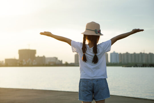 Asian kid girl wearing hat raising arms relaxing feeling happy looking at sunset on sky enjoying sunlight and Thatluang lake view in Vientiane city, Laos during vacation, freedom hope concept.