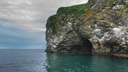 Fototapeta premium An island in the Pacific Ocean. Seabirds nest on steep rocky slopes with sparse vegetation. The entrance to the dark grotto is visible above the surface of the water. Clouds in the sky. Kamchatka.