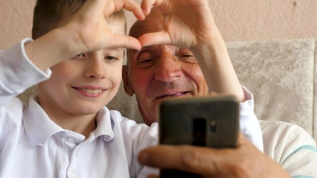 cheerful caucasian boy 7 years old and pensioner 70 years old posing in front of phone camera taking selfie and showing heart shaped gesture. grandson and grandfather home life
