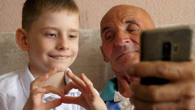 
Caucasian boy and grandfather 70 years old posing in front of smartphone camera taking selfie at home.Child showing heart shaped gesture.Slow motion