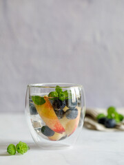 Blueberry and peach infused water, cocktail, lemonade or tea in double glass. Summer iced cold drink with blueberry, ice, peach and mint on grey marble background, copy space, vertical, side view
