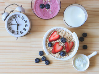 flat lay of alarm clock, breakfast with oat or granola in white bowl, fresh blueberries, strawberries, a  glass of milk, blueberry smoothie  and yogurt on wooden table. Healthy breakfast concept.