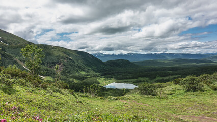 Fototapeta na wymiar View of the lake from the mountain slope. The meadow has lush green vegetation. The path goes down. Picturesque clouds in the blue sky. Reflection. Kamchatka. Vachkazhets. Lake Tahkoloch