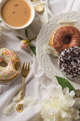 Top view of breakfast in bed with various delicious glazed donuts on the plate, cup of coffee and fresh peony flowers.