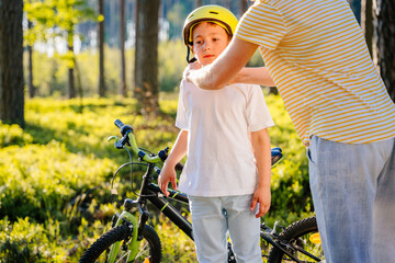 Unrecognizable mother helping son wearing helmet for cycling at forest. Child getting ready by wearing bike helmet to start cycling.