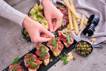 woman puts arugula leaves to prosciutto ham bruschetta with traditional antipasto meat plate on background