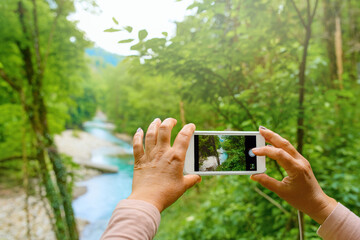 A woman photographs nature with a cell phone. The phone in photo mode is aimed at a mountain river.