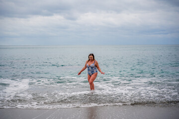A plump woman in a bathing suit enters the water during the surf. Alone on the beach, Gray sky in the clouds, swimming in winter.
