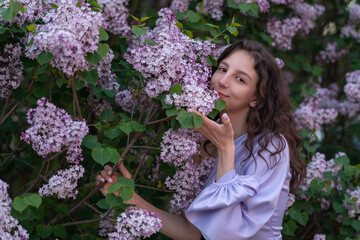 Portrait of a beautiful girl in a lilac dress is standing near a bush breathing the aroma of lilacs