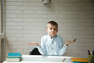 A boy of 6-7 years old at a white desk shows a don't know gesture against a brick wall. School education - obrazy, fototapety, plakaty
