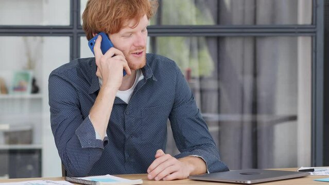 Concentrated redhead businessman office worker opens laptop and starts working talking on a mobile phone with partners or employees, discussing something sitting in a modern office or coworking