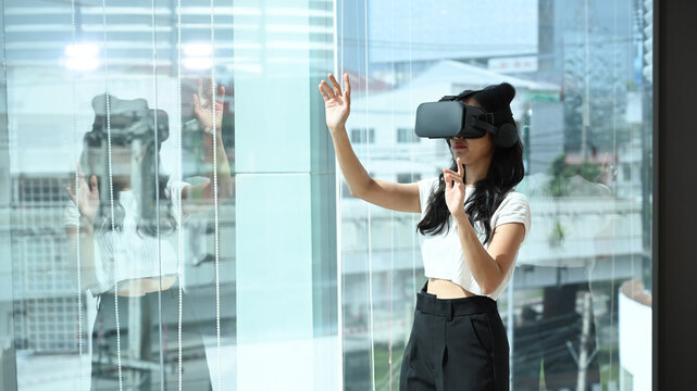 Young woman wearing virtual reality headset and interacts with cyberspace, standing near large window with city buildings view outside