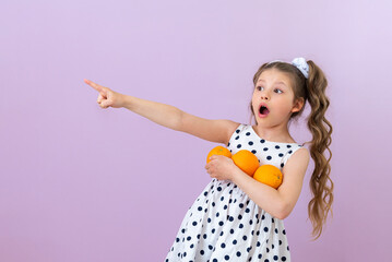 A little girl holds oranges and points to an advertisement on an isolated pink background.