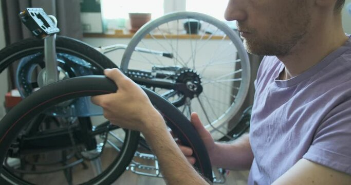 man holds a bicycle camera inner tube in his hands, shows taped punctures on it. Home bicycle workshop with a master. Close-up, disassembled bike in the background.