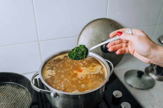 A woman, a girl, a cook prepares vegetable soup in boiling water, pouring green dill from a spoon into a saucepan. Cooking food in the kitchen.