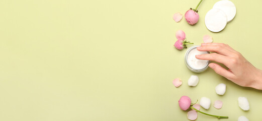 Female hand with jar of cosmetic cream, cotton balls, pads and rose flowers on green background with space for text