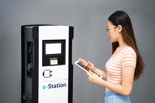 Young Asian Thai, Vietnamese Or Chinese Woman In Casual Clothes And Glasses Holding Tablet, Using Touchscreen Electric Charge Station Near Modern Car And Office Building Outside. Car Sharing, Energy