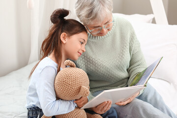Little girl with toy and her grandma reading book in bedroom