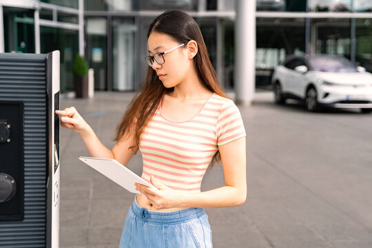 Young Asian Thai, Vietnamese or Chinese woman in casual clothes and glasses holding tablet, using touchscreen electric charge station near modern car and office building outside. Car sharing, energy