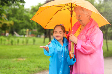 Little girl and her grandma in raincoats with umbrella outdoors