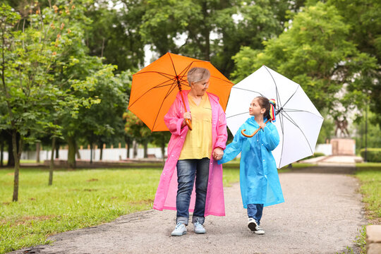Little girl and her grandma in raincoats with umbrellas walking outdoors