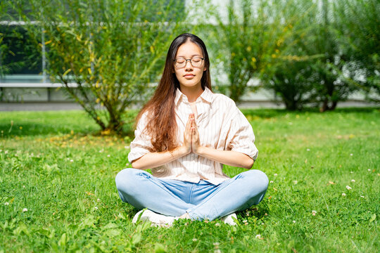 Young Asian Thai, Vietnamese or Chinese woman in casual clothes sitting in lotus yoga pose meditating on grass loan near bushes and glass office building on weekend after work on sunny summer day. 