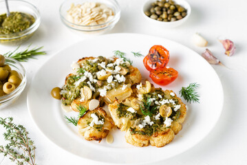 Cauliflower steak with spices, chimichurri sauce, almond flakes, olives, fried cherry tomatoes and capers on a white plate. Vegetarian food.