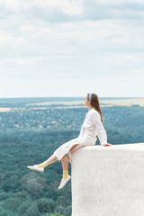 Fototapeta na wymiar Young woman in white clothes sits on high observation deck with her legs hanging down. Full-length portrait. Vertical frame.